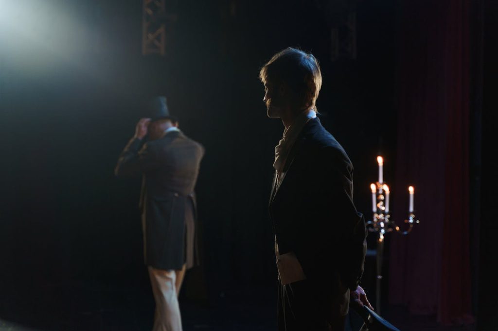 Two actors in Victorian costumes perform a dramatic scene on stage, lit by candlelight.